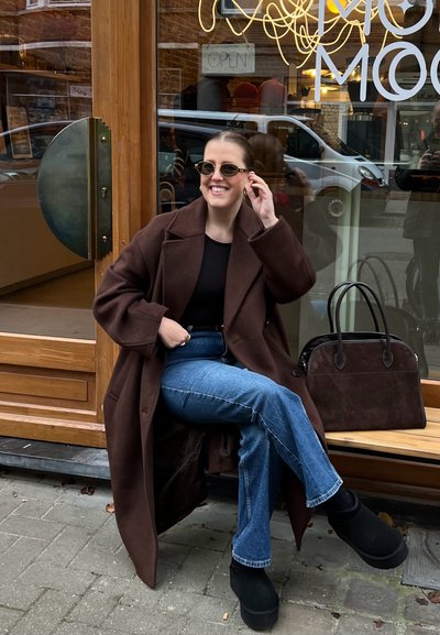 Abrigo de lana marrón sobre una blusa negra, combinado con jeans azul de corte acampanado y botas de plataforma negra. Un bolso oscuro descansa a su lado.