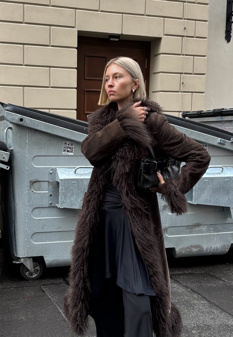 Woman with short blonde hair wearing a long dark fur-lined coat and holding a small black bag, standing near metal dumpsters.