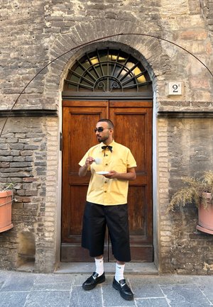 Camisa amarilla de manga corta con botones, pantalones cortos negros hasta la rodilla, mocasines negros, sosteniendo una taza y un plato blancos, de pie junto a una puerta de madera.