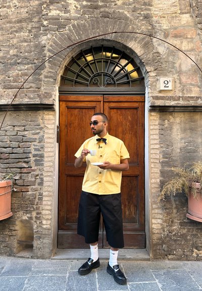 Camisa amarilla de manga corta con botones, pantalones cortos negros hasta la rodilla, mocasines negros, sosteniendo una taza y un plato blancos, de pie junto a una puerta de madera.