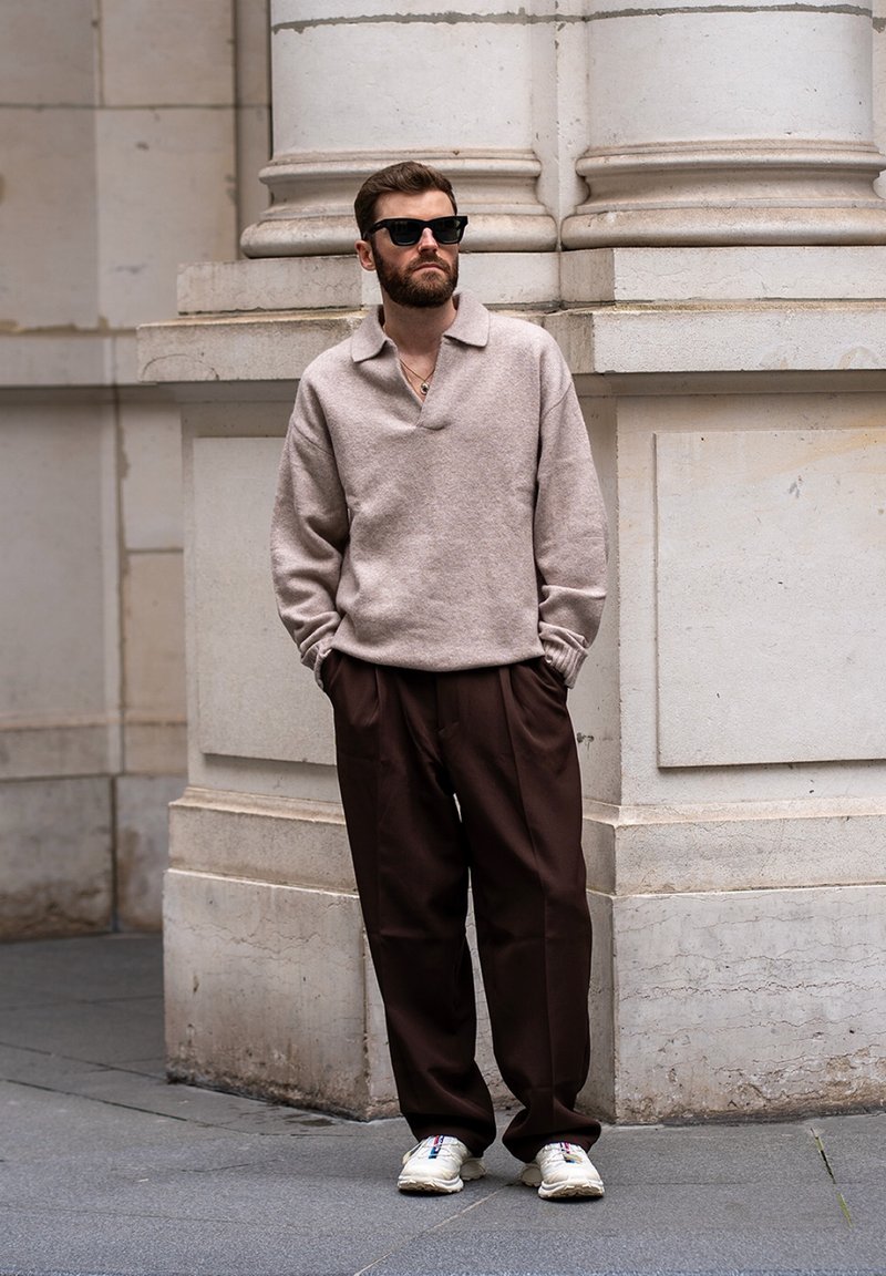 Homme portant un pull beige, un pantalon marron foncé, des baskets blanches et des lunettes de soleil noires, debout les mains dans les poches près d'une colonne en pierre en plein air.
