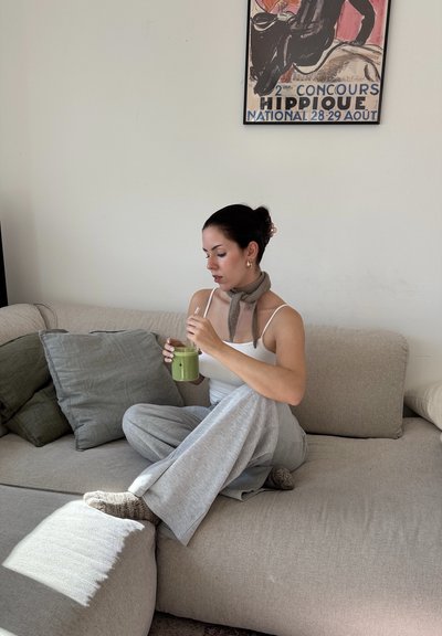 Un sofá de tela gris claro con cojines texturizados en varios tonos de verde. Una persona con una camiseta sin mangas sostiene una bebida verde y está sentada con las piernas cruzadas.