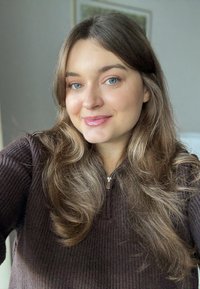 Młoda osoba dorosła z długimi, falowanymi brązowymi włosami, ubrana w ciemny prążkowany sweter z zamkiem błyskawicznym. Tło cechuje się delikatnymi, neutralnymi kolorami. Naturalne oświetlenie.