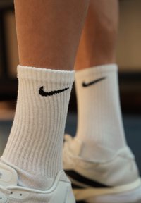 Hvite Nike-sokker brukt med hvite treningssko på en person som står innendørs på en myk overflate.
