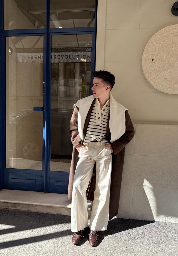 Jonge man die een lange bruine jas draagt, een gestreepte trui, crèmekleurige wijde broek en bruine schoenen, staand bij een blauwe deur met de tekst "Franse Revolutie".