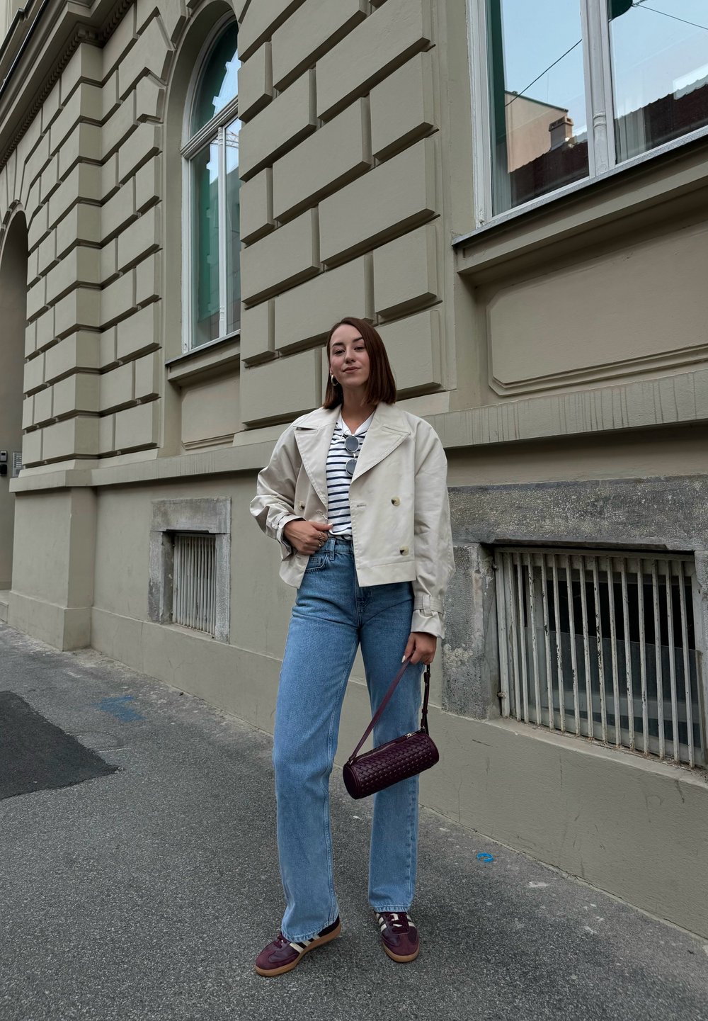 Jachetă cropped bej deschis, cămașă cu dungi, blugi drepti albaștri și geantă adânc purpurie cu un model texturat. Tenisi cu accente întunecate.