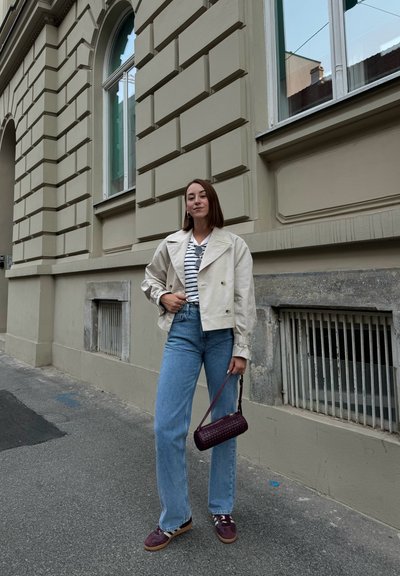 Chaqueta cropped en beige claro, camisa a rayas, jeans rectos azules y bolso de mano púrpura oscuro con un patrón texturizado. Zapatillas con detalles oscuros.