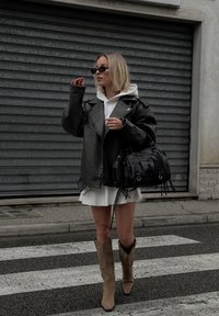Černá kožená motorkářská bunda, bílá mikina, bílé šaty, béžové kovbojské boty a černá kabelka s kovovými akcenty. Modelka stojí na ulici.