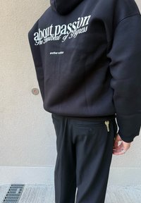 Persoană îmbrăcată într-un hanorac negru cu text alb "despre pasiune" și pantaloni negri, având o cheie în buzunarul din spate, stând lângă un zid.