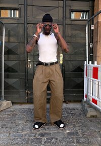 Balta liemenė, laisvi chaki spalvos kelnių, juodas diržas, juodi šlepetės su baltais padais ir saulės akinių. Stovi prieš tamsiai medinį foną.