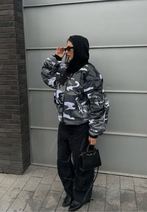 Chaqueta bomber de camuflaje gris y blanco con puños acanalados, combinada con pantalones negros y un pequeño bolso negro. Modelo llevando gafas de sol y capucha.