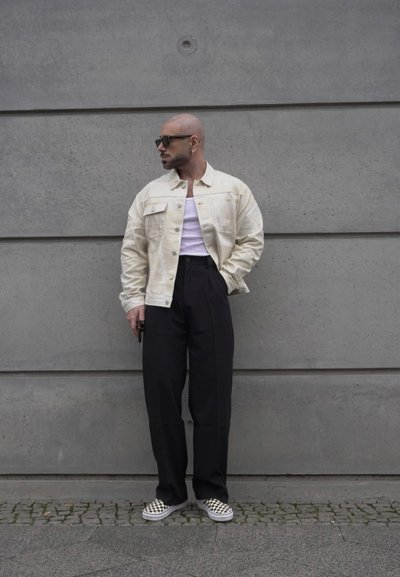 Chaqueta de mezclilla crema, camiseta blanca sin mangas, pantalones negros de tiro alto y zapatos de lona a cuadros. Textura suave contra un fondo de hormigón.