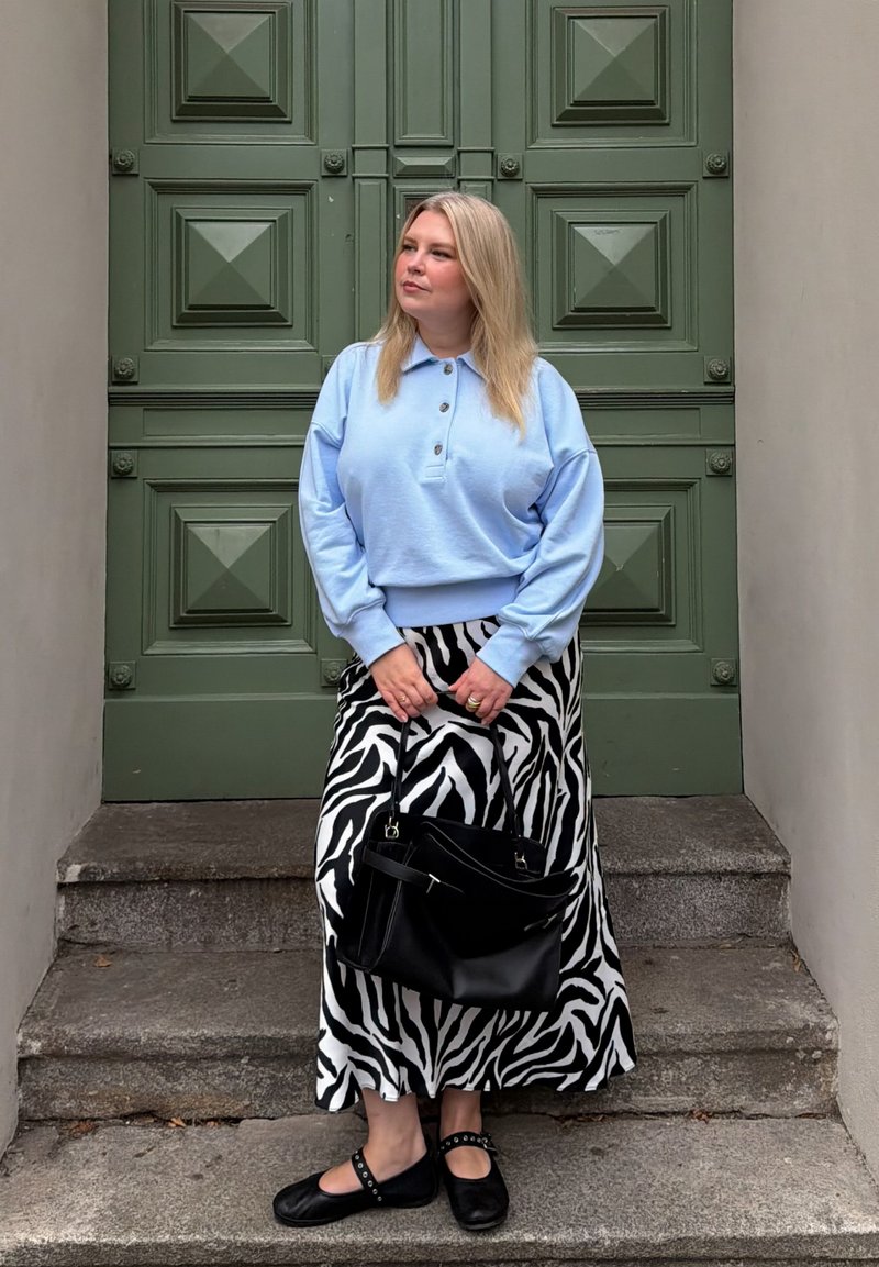 Woman with blonde hair wearing a light blue sweater and zebra print skirt, holding a black handbag, standing on stone steps.