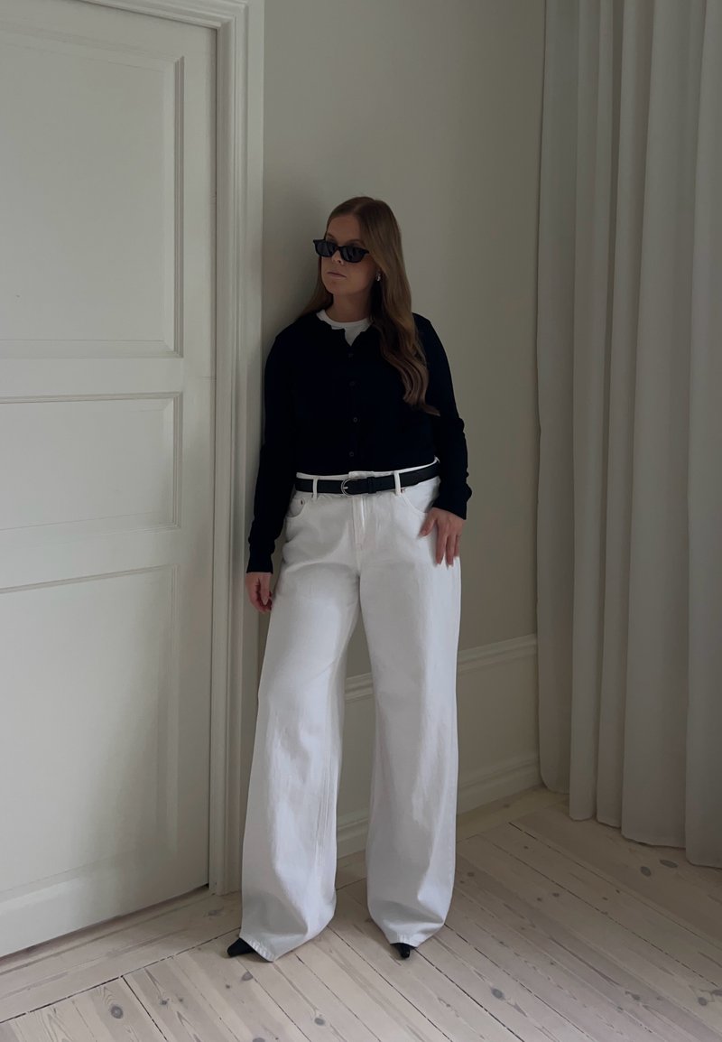 Femme portant un haut noir à manches longues, un pantalon blanc à jambes larges, une ceinture noire et des lunettes de soleil noires, debout à l'intérieur près d'une porte et d'un rideau blanc.