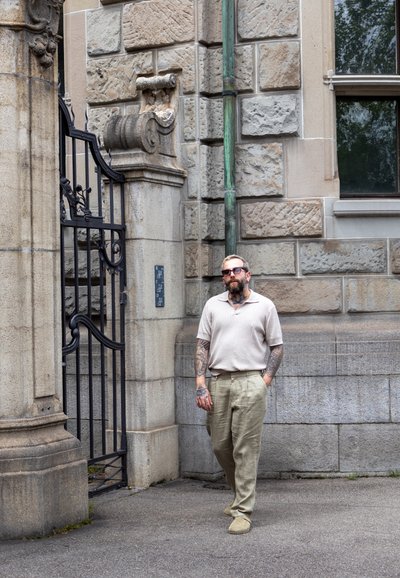 Camisa polo de canalé beige claro combinada con pantalones de lino caqui claro. De pie cerca de una arquitectura de piedra con detalles intrincados y una puerta de metal.