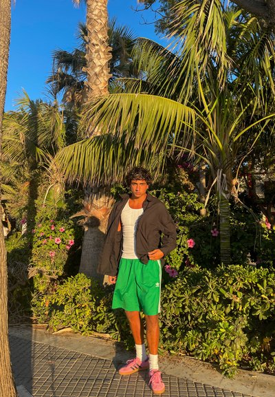 Una persona se encuentra frente a palmeras, vistiendo una chaqueta negra, una camiseta blanca de tirantes, pantalones cortos verdes, zapatos rosas y calcetines blancos.