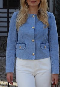 Vaaleansininen farkkutakki, jossa on pyöreä kaulus, kultaiset napit ja kaksi etutaskua. Yhdistetty valkoisiin housuihin.