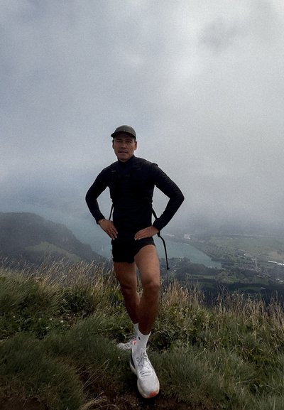 Atleta con una camiseta de manga larga negra y pantalones cortos negros, de pie en la hierba con una vista de colinas y un cuerpo de agua de fondo.