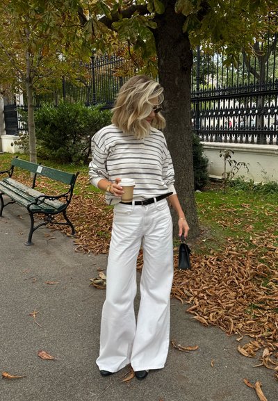 Suéter a rayas gris claro, pantalones anchos blancos, cinturón negro, sosteniendo una taza de café. Bolso negro, hojas caídas y un banco del parque al fondo.