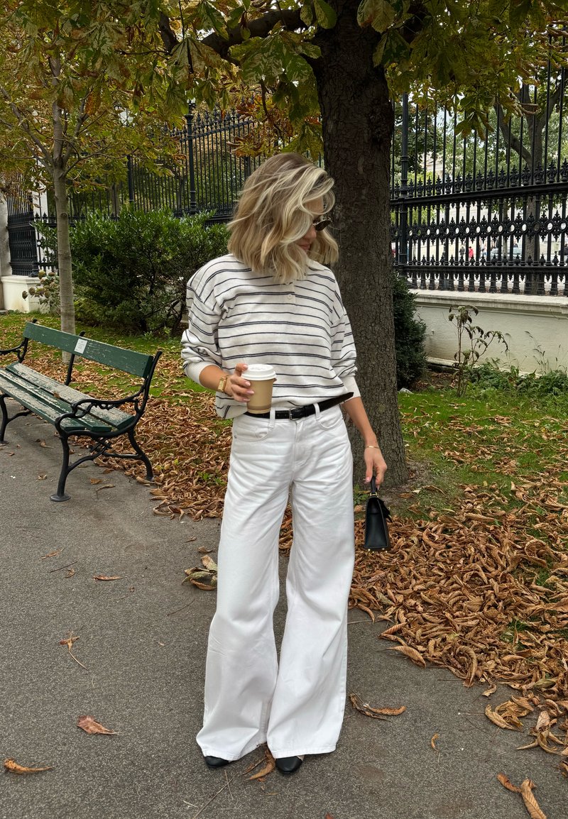 Striped light grey sweater, white wide-leg trousers, black belt, holding a coffee cup. Black handbag, fallen leaves, and a park bench in the background.