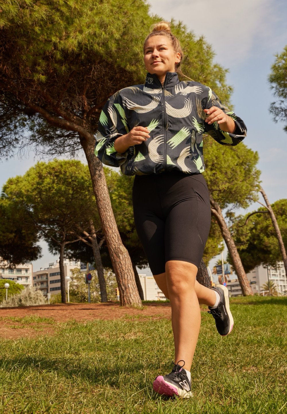 Veste coupe-vent légère à motifs, de couleur bleu foncé avec des accents verts et blancs, associée à un short de vélo noir et des chaussures de course avec des détails roses.