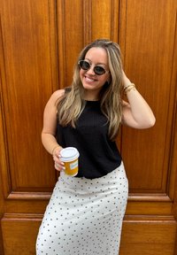 Schwarzes, strukturiertes ärmelloses Top, Rock mit weißen Punkten, hält eine weiße Kaffeetasse mit gelbem Etikett, trägt runde schwarze Sonnenbrille.