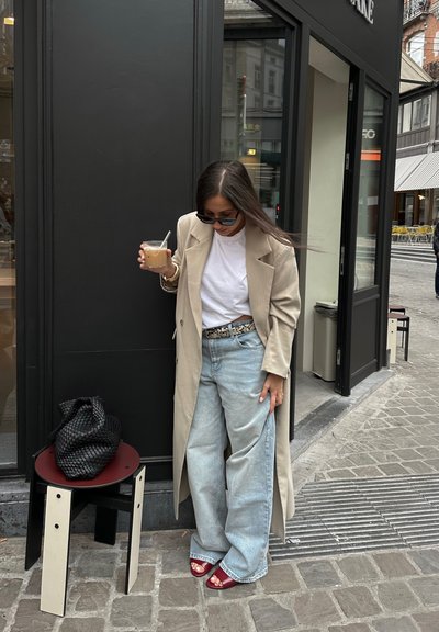 Abrigo largo beige sobre camiseta blanca, jeans de pierna ancha azul claro, sandalias rojas de punta abierta. Sostenido una bebida, con un bolso de mimbre negro sobre un taburete.
