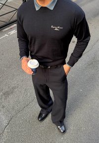 Pulover lung negru cu mâneci, purtat peste o cămașă cu guler de culoare albastru deschis, asortat cu pantaloni negri și pantofi din piele. Ține o cană de cafea.