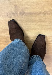 Pruun sametisest nahast küberkummikud terava varbaga, dekoratiivse õmbluse ja siledapõhjaga, kantud siniste teksapükstega.