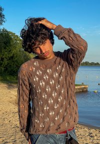 Pull en tricot marron avec un motif de feuilles, présentant un design ample et léger. En arrière-plan, on trouve de l'eau et des arbres sous un ciel bleu dégagé.