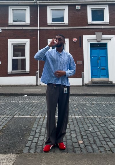 Hombre con una camisa de rayas azul y blanca y pantalones grises, sosteniendo una bebida. Lleva zapatos rojos y una placa. Fondo de una calle empedrada y un edificio de ladrillo.