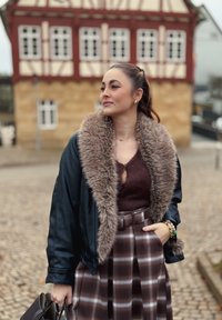 Veste en cuir noir avec un col en fausse fourrure, haut en tricot marron et jupe à carreaux dans des tons bruns. Les accessoires comprennent un sac à main et des bracelets.