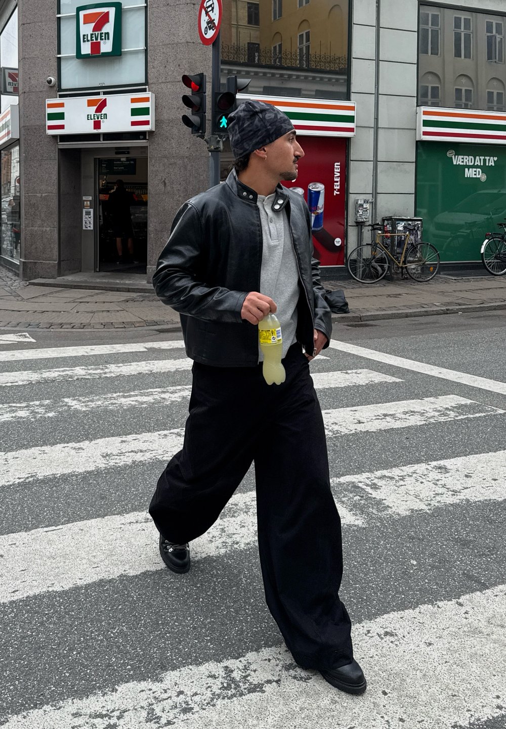 Személy gyalogosátkelőhelyen, fekete bőrkabátot, szürke inget és bő, fekete nadrágot viselve. Sárga italt tart a kezében. Városi háttér.