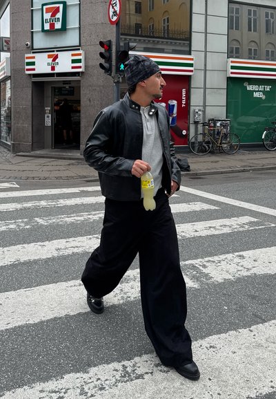 Persona cruzando un paso de peatones, vistiendo una chaqueta de cuero negra, camisa gris y pantalones negros sueltos. Sosteniendo una bebida amarilla. Fondo urbano.