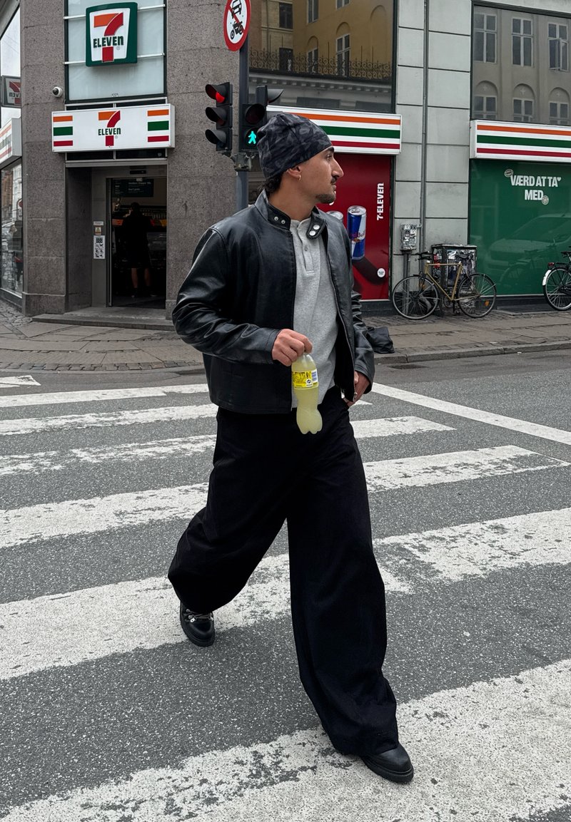 Henkilö kävelee suojatiellä yllään musta nahkatakki, harmaa paita ja väljät mustat housut. Hän pitelee keltaista juomaa. Kaupunkimaisema taustalla.