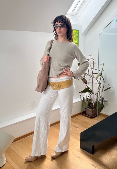 Suéter gris claro, pantalones blancos de campana, cinturón decorativo amarillo con tachuelas, bolso marrón de hombro y tacones transparentes. Cabello rizado, gafas de sol.