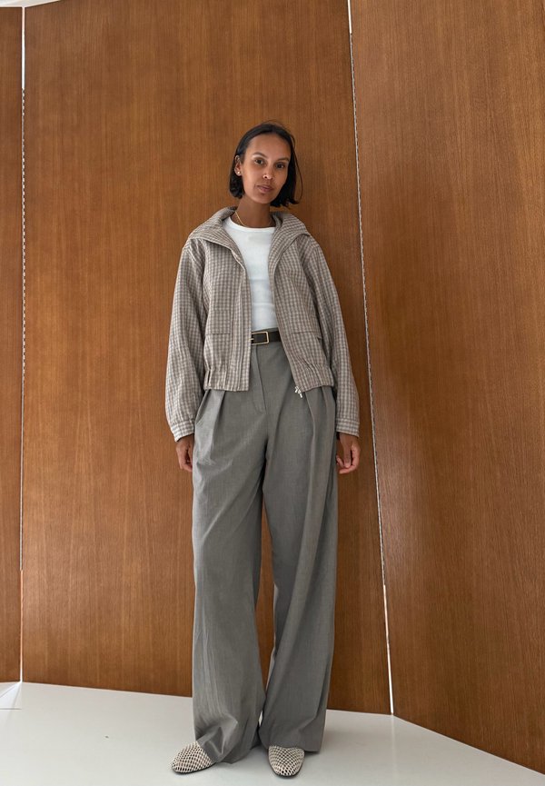 Mujer de pie junto a paneles de madera, vestida con una camiseta blanca, chaqueta beige de cuadros, pantalones anchos grises y zapatos planos con estampado.