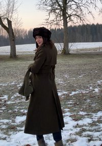 Γυναίκα με μακρύ σκοτεινό παλτό και γούνινο καπέλο, που στέκεται σε χιονισμένο χωράφι με γυμνά δέντρα στο λυκόφως, κοιτώντας πίσω πάνω από τον ώμο της.