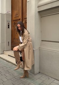 Manteau beige trench avec des accents de boutons ; modèle ajustant des bottes marron montantes avec une finition texturée ; trottoir et porte en bois en arrière-plan.