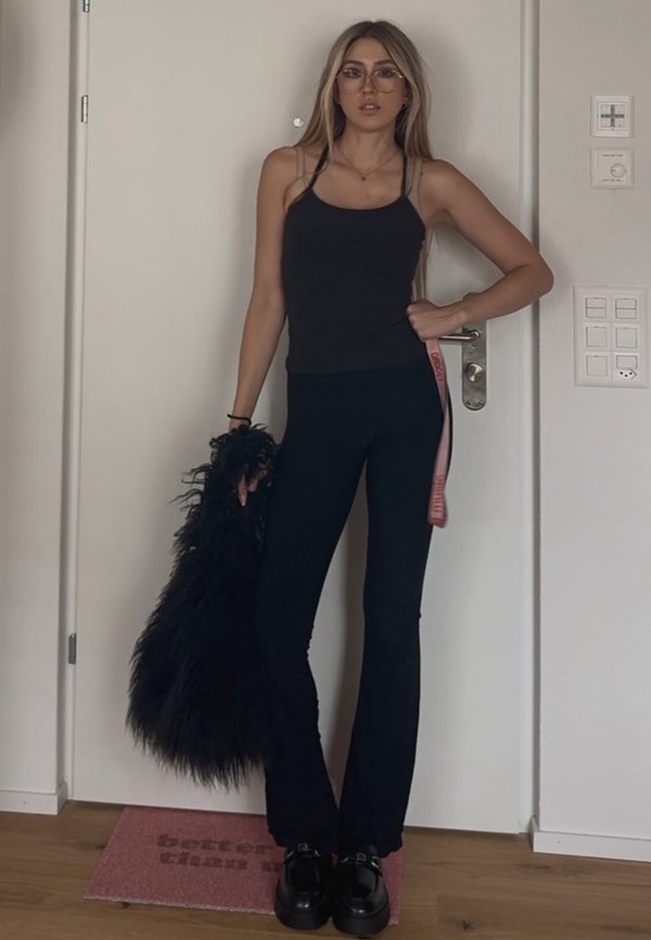 Mujer con cabello largo y rubio y gafas lleva una camiseta sin mangas negra y pantalones, sosteniendo una gran bolsa negra y peluda, de pie en el interior junto a una puerta.