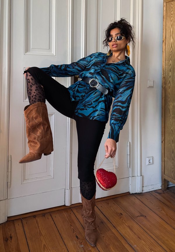 Femme posant à l'intérieur portant un chemisier bleu à motifs, un pantalon noir, des collants imprimé léopard, des bottes marron, des lunettes de soleil, tenant un sac rouge en forme de cœur.