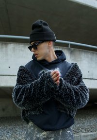 Veste en fausse fourrure noire avec des manches larges, superposée à un sweat à capuche noir. Portée avec des lunettes de soleil sombres et un bonnet noir. Texturé et contemporain.