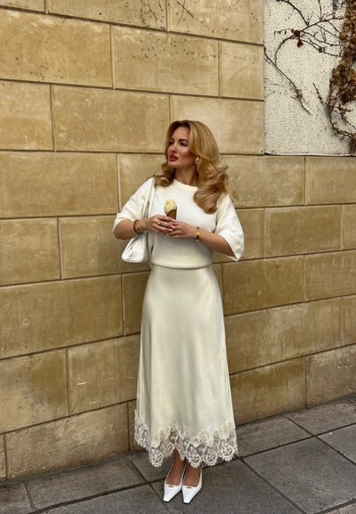 Mujer con vestido blanco y suéter sosteniendo un cucurucho de helado de vainilla, frente a una pared beige con azulejos y enredaderas a un lado.