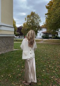 Cardigan crème avec des boutons dans le dos, associé à une longue jupe beige en soie. Texture douce et coupe décontractée sur un fond herbeux.