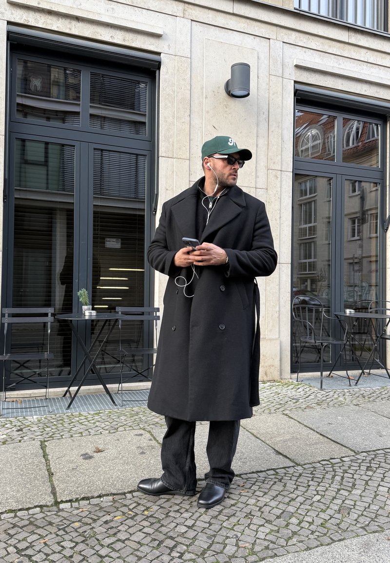 Schwarzer, doppelt geknöpfter Mantel, schwarze Jeans und Schuhe. Trägt eine Sonnenbrille und eine dunkelgrüne Mütze. Hält ein Smartphone mit Ohrhörern. Draußen.