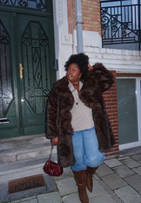 Γυναίκα με σκούρο γούνινο παλτό, μπεζ τοπ, μπλε τζιν και καφέ μποτάκια στέκεται σε πέτρινο πλακόστρωτο κοντά σε πράσινη πόρτα, κρατώντας μια μικρή κόκκινη τσάντα χειρός.