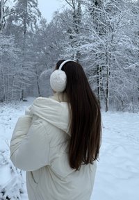 Casaco puffer branco com um grande colarinho, lenço creme e protetores de ouvidos de pelo sintético brancos. Cabelo castanho comprido em frente a um fundo de floresta nevada.