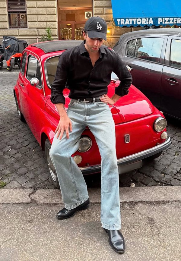 Röd vintagebil med runda strålkastare parkerad på kullersten. Person i svart skjorta, ljusblå jeans och svarta stövlar poserar bredvid den.