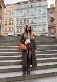 Brauner knieumspielender Mantel über weißem Oberteil und braunen Shorts. Braune Handtasche mit kurzem Griff. Lange braune Stiefel. Steinige Treppe im Hintergrund.