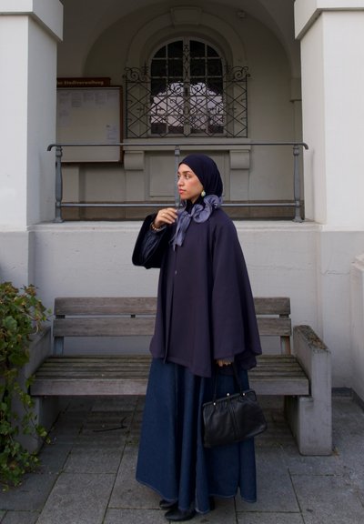 Capa azul marino sobre una larga falda de mezclilla, combinada con un bolso oscuro. Presenta una bufanda azul clara y pendientes sutiles. Fondo urbano.